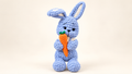 Blue handmade crochet bunny holding an orange carrot on a cream background