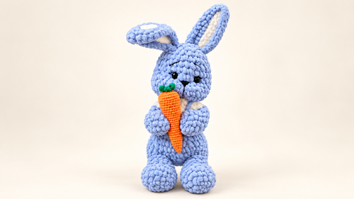 Blue handmade crochet bunny holding an orange carrot on a cream background